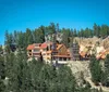 An expansive lodge-style building perched on a forested hillside with rocky cliffs under a clear blue sky