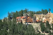 An expansive lodge-style building perched on a forested hillside with rocky cliffs under a clear blue sky.