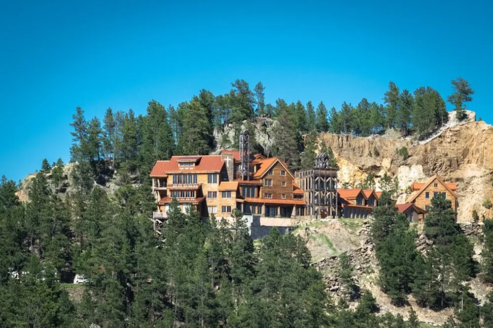 An expansive lodge-style building perched on a forested hillside with rocky cliffs under a clear blue sky