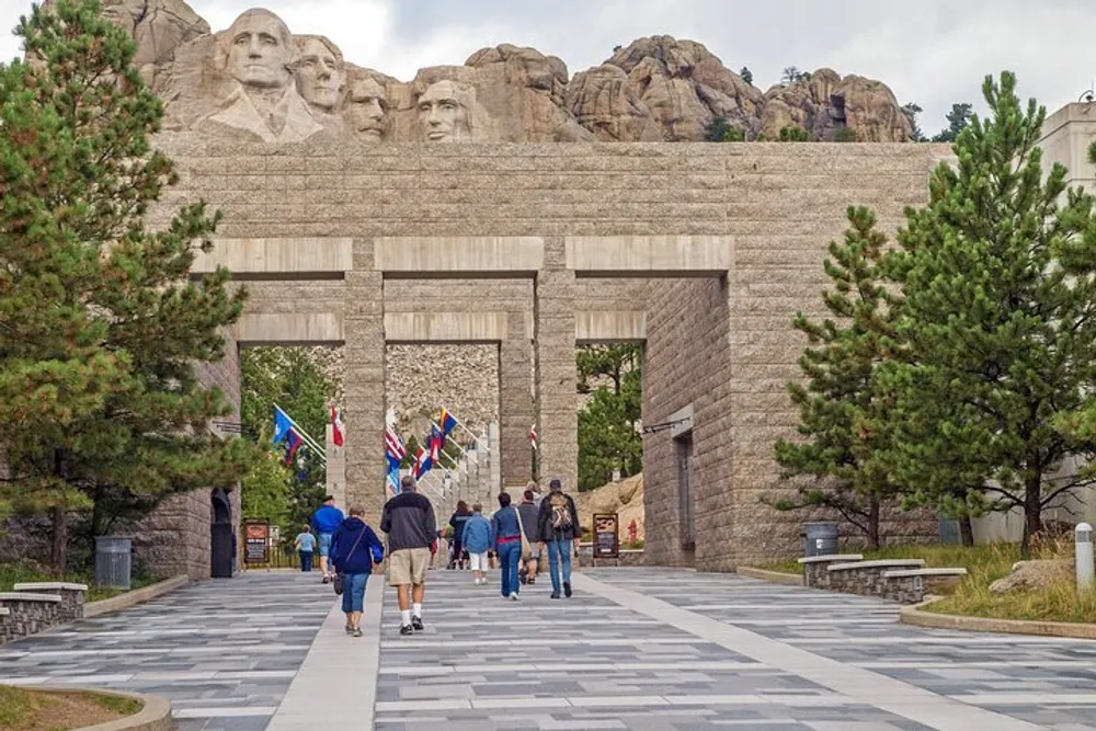 Visitors walk towards the entrance of a monument with the iconic Mount Rushmore National Memorial featuring four presidential faces carved into the mountain visible in the background