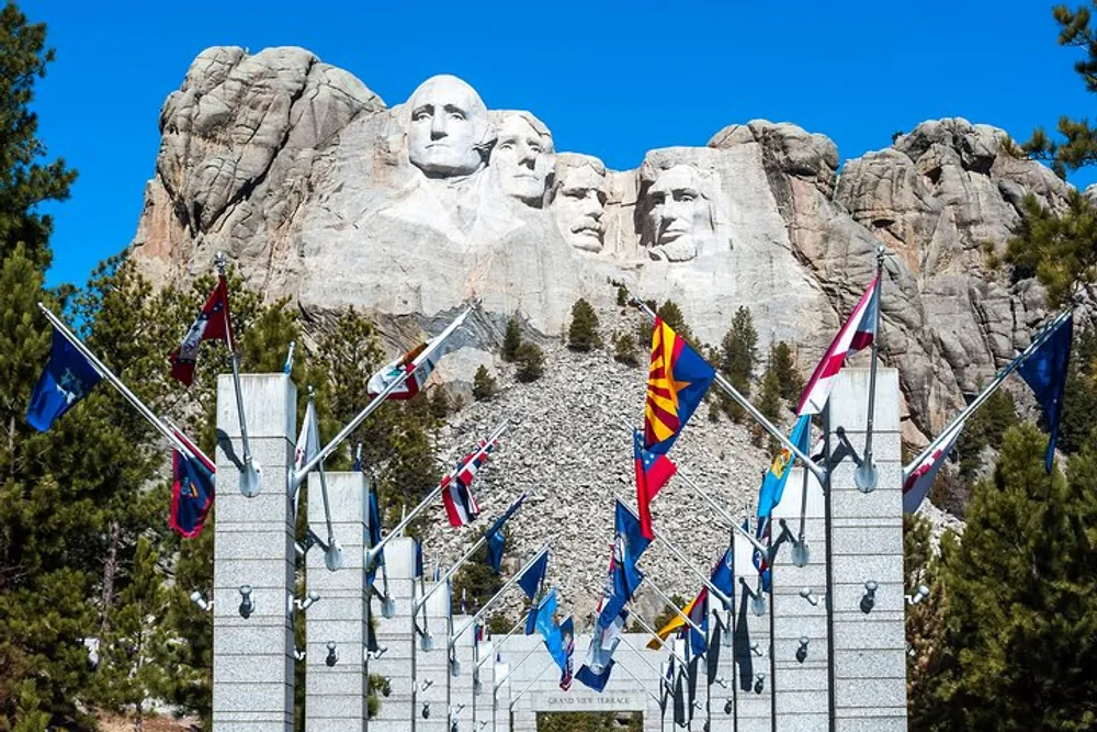 The image showcases Mount Rushmore a massive sculpture carved into the Black Hills of South Dakota with a clear blue sky backdrop and an avenue of United States flags leading up to it