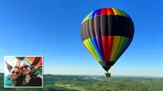 A colorful hot air balloon is soaring over a landscape with a small inset of a smiling couple taking a selfie inside it.