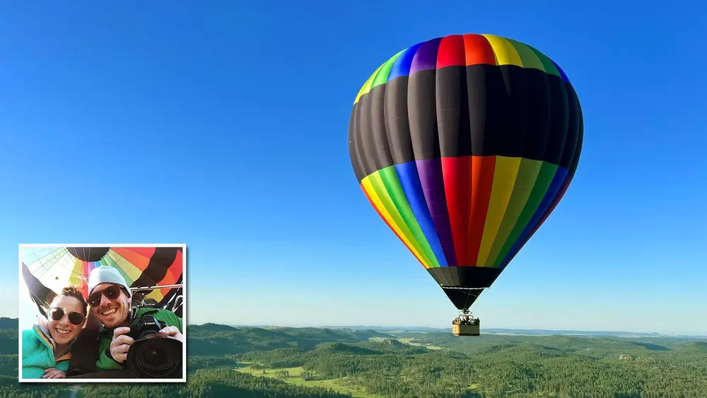 A colorful hot air balloon is soaring over a landscape with a small inset of a smiling couple taking a selfie inside it