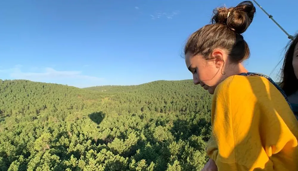 A person in a yellow shirt is looking down over a lush green forest with a hot air balloon shadow visible on the trees