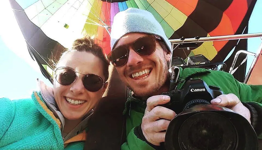 A smiling couple wearing sunglasses takes a selfie with a camera with a colorful hot air balloon visible in the background