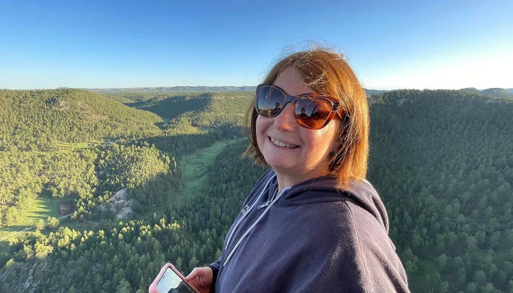 A woman with sunglasses smiles while holding a phone standing against a backdrop of a vast green landscape under a clear blue sky