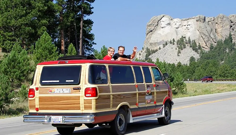 Two people are cheerfully popping out of the roof of a classic wood-paneled van with Mount Rushmore in the background