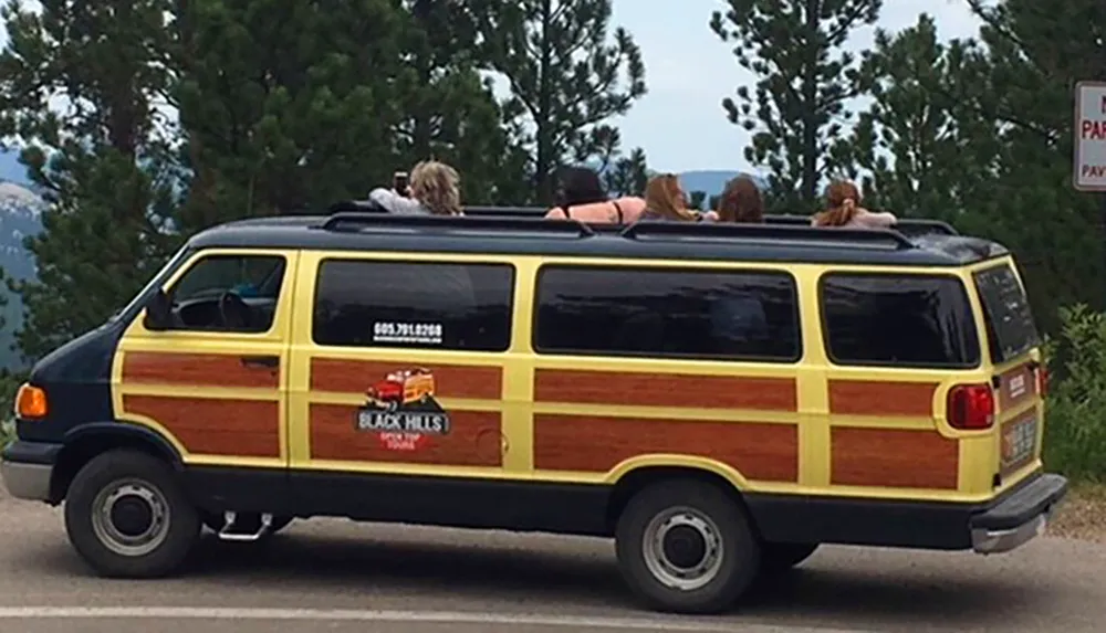 A group of people is lying on the roof of a moving van that is decorated to look like a wood-paneled vehicle with the words Black Hills Tours on the side