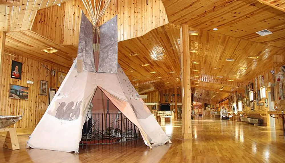 The image shows a large indoor space with wooden walls and flooring featuring a traditional tipi at its center surrounded by various cultural exhibits and artifacts