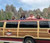 A group of people is posing on the roof of a wood-paneled van with craggy rock formations in the background
