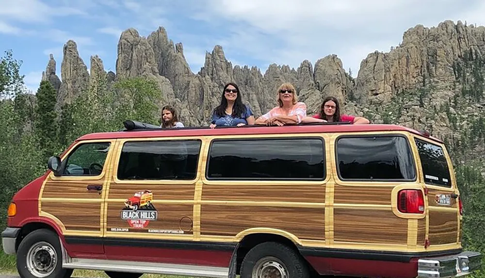 A group of people is posing on the roof of a wood-paneled van with craggy rock formations in the background