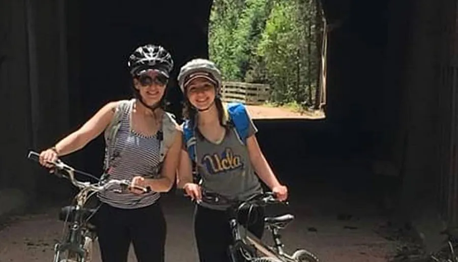 Two smiling people with bicycles are standing in front of a dark tunnel, wearing helmets and casual clothing for a bike ride.