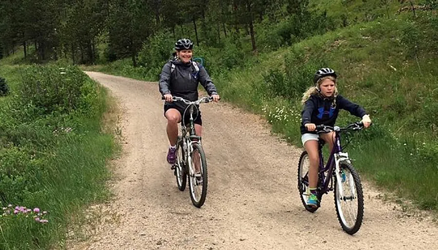 Two people are enjoying a bike ride on a dirt path through a green, wooded area.