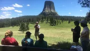 A group of people is listening to a guide who is giving a presentation in front of the impressive Devils Tower, a notable geological feature that protrudes out of the rolling prairie in Wyoming.