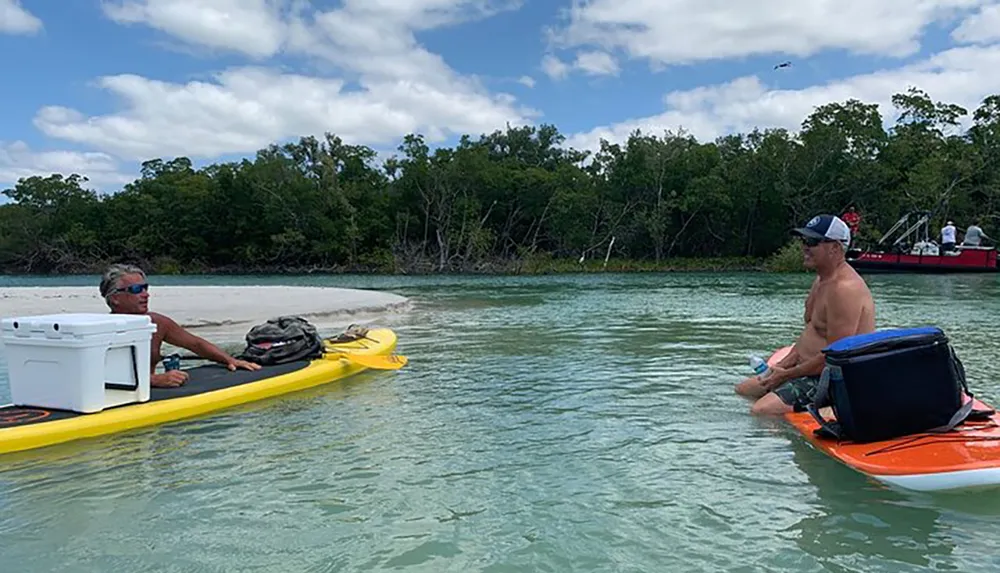 Two people are enjoying a sunny day on the water with one sitting on a yellow kayak and the other on an orange paddleboard surrounded by clear waters and greenery