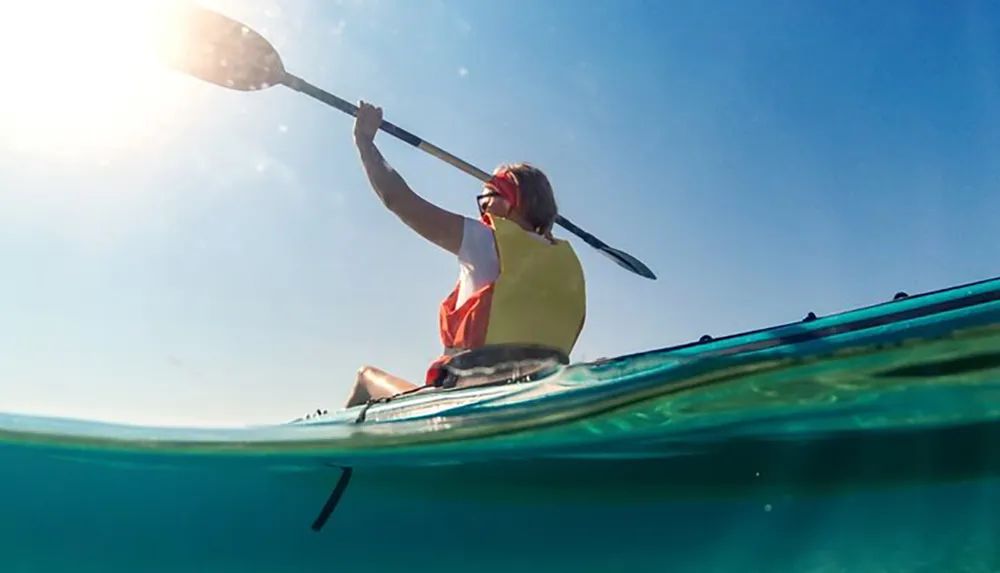 A person is kayaking in clear blue waters captured from an underwater perspective that splits the image between the above-water scenery and the sub-surface environment