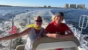Two people are smiling and enjoying a sunny boat ride with a view of the water and buildings in the distance.