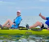 Two people are smiling and giving a thumbs-up while sitting on a tandem kayak in calm water