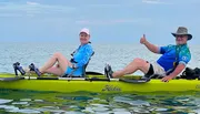 Two people are smiling and giving a thumbs-up while sitting on a tandem kayak in calm water.