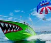 A brightly colored speedboat with a shark face design is towing two people on a parasail above a clear blue sea