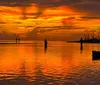 The image captures a serene sunset with vibrant orange hues reflecting on the waters surface where silhouetted individuals are engaged in fishing activities
