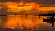 The image captures a serene sunset with vibrant orange hues reflecting on the water's surface, where silhouetted individuals are engaged in fishing activities.