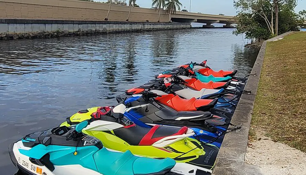 A row of colorful personal watercraft is parked along the edge of a calm waterway near a bridge