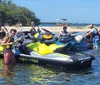 A group of people wearing life vests are posing with their personal watercraft on a sunny day near the shore