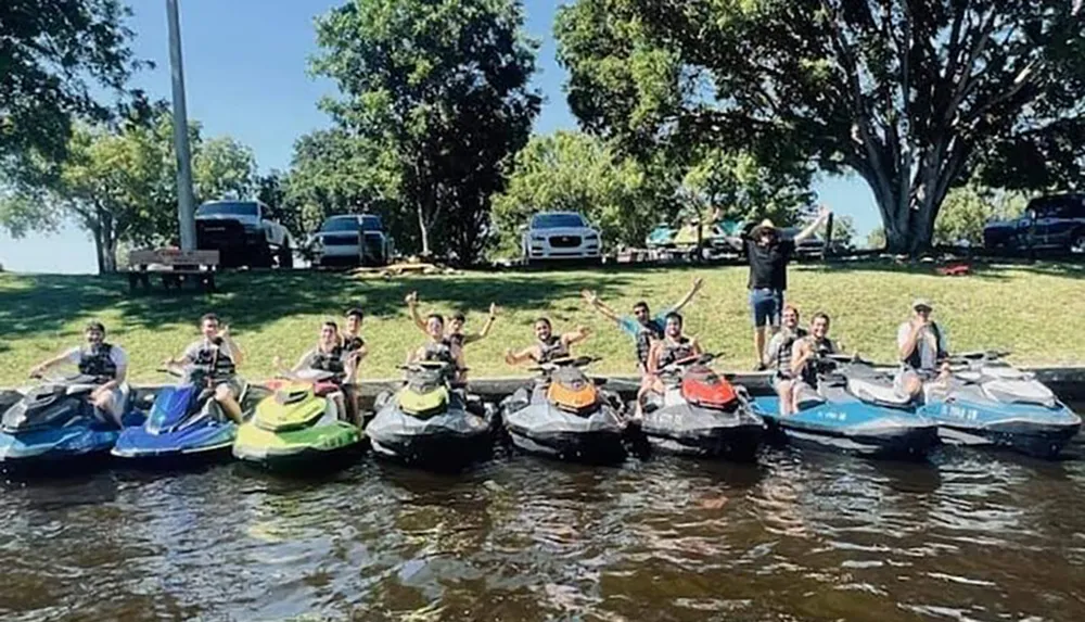 A group of people is posing cheerfully on personal watercraft at the edge of a sunlit body of water with trees and vehicles visible in the background