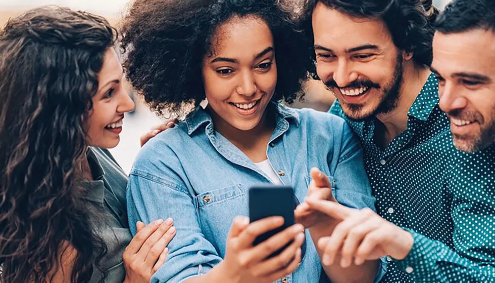 A group of four smiling friends are closely gathered and looking at a smartphone together