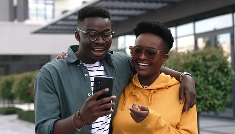 Two people are smiling and looking at a smartphone together with a backdrop of urban architecture