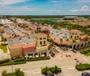 The image shows an aerial view of a sunny suburban shopping center with terracotta-roofed buildings and parked cars indicating a bustling commercial area