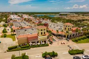 The image shows an aerial view of a sunny suburban shopping center with terracotta-roofed buildings and parked cars, indicating a bustling commercial area.