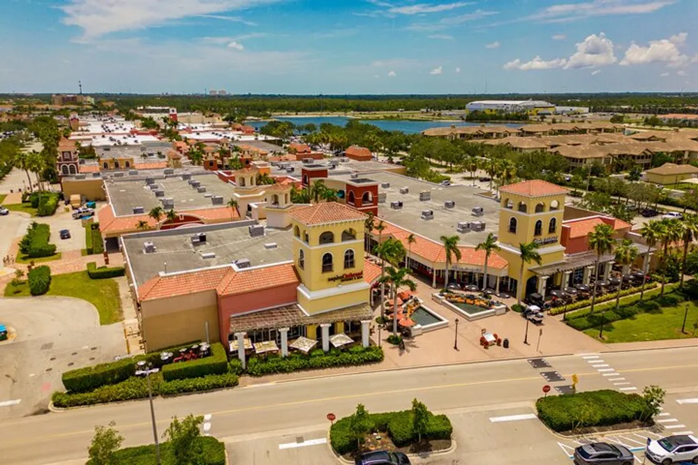 The image shows an aerial view of a sunny suburban shopping center with terracotta-roofed buildings and parked cars indicating a bustling commercial area