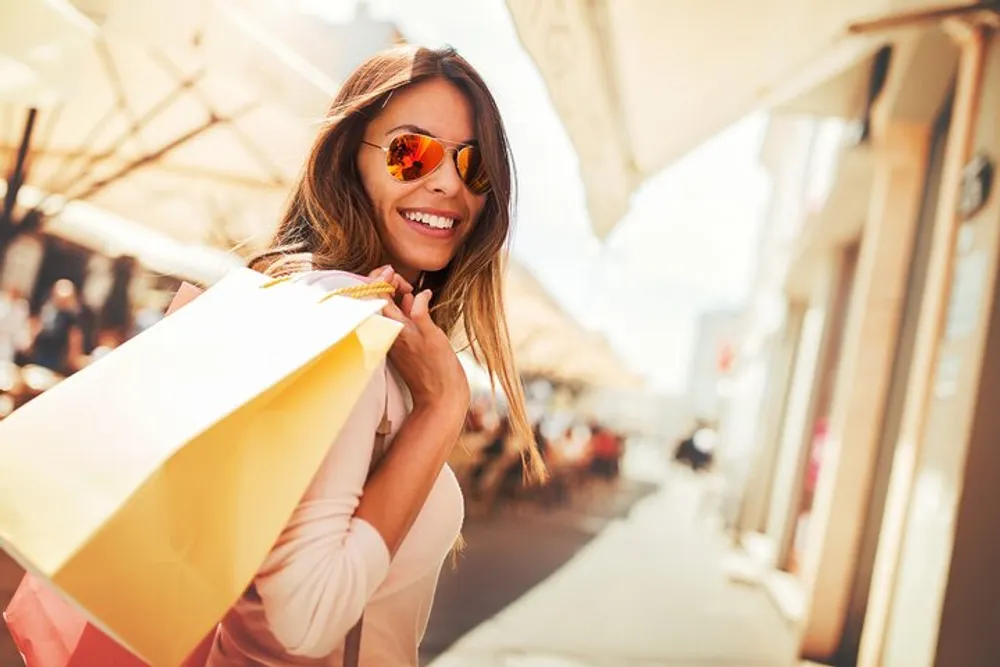 A smiling woman wearing sunglasses is holding shopping bags on a sunny street with storefronts and pedestrians in the background