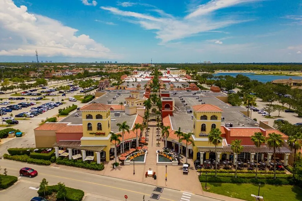 The image shows an aerial view of a sunny palm-lined shopping plaza with terracotta roofs bustling with activity surrounded by an expansive parking lot