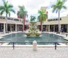 The image shows a sunny outdoor shopping area with a central fountain flanked by palm trees and storefronts including US Polo Assn and Converse with people casually walking by