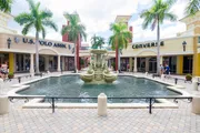 The image shows a sunny outdoor shopping area with a central fountain, flanked by palm trees and storefronts including U.S. Polo Assn. and Converse, with people casually walking by.