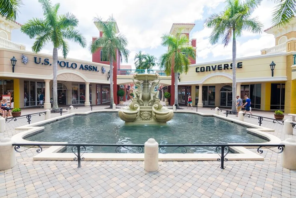 The image shows a sunny outdoor shopping area with a central fountain flanked by palm trees and storefronts including US Polo Assn and Converse with people casually walking by