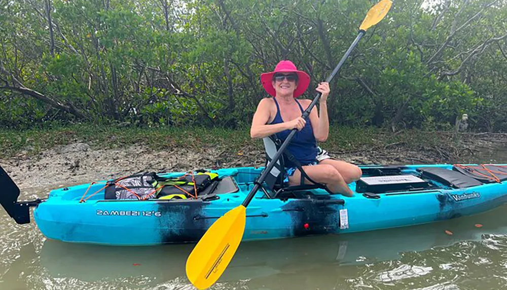 A person is happily kayaking in a blue kayak holding a yellow paddle and wearing a bright red hat