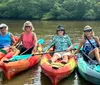 Four people are smiling for the camera while seated in colorful kayaks on calm waters with lush greenery in the background