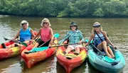 Four people are smiling for the camera while seated in colorful kayaks on calm waters, with lush greenery in the background.