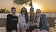 Four friends are enjoying a sunset together on a beach, wearing flower necklaces and holding drinks.