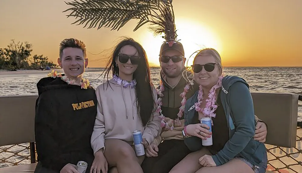 Four friends are enjoying a sunset together on a beach wearing flower necklaces and holding drinks