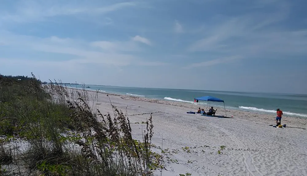 This image shows a tranquil beach scene with people relaxing under a blue canopy and a clear sky