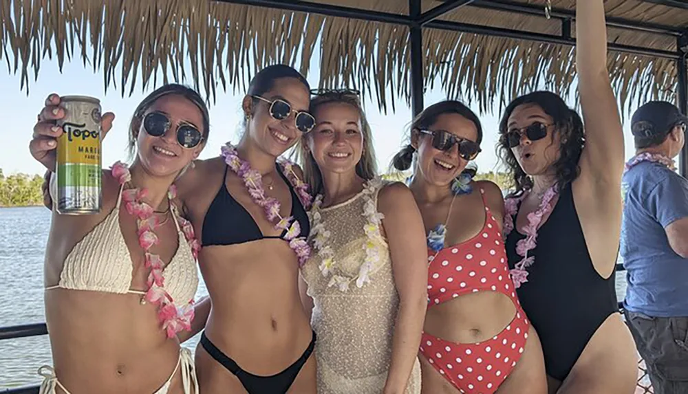 Five women wearing sunglasses and floral leis are smiling and posing for a photo together with one holding up a beverage can while under a thatched roof structure suggesting a tropical or festive setting