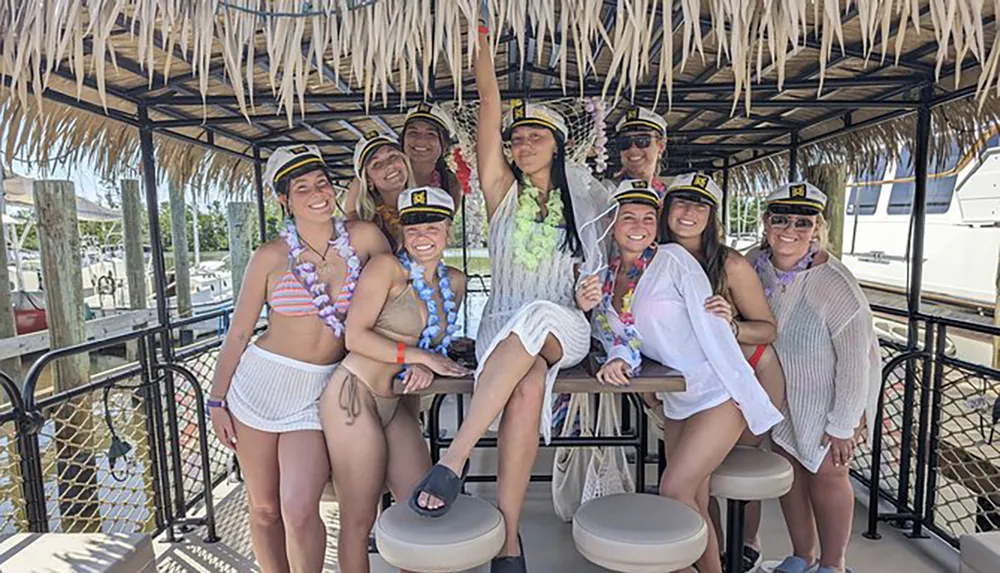 A group of people is posing joyfully under a thatched roof structure some wearing captains hats and lei necklaces suggesting a festive or celebratory occasion