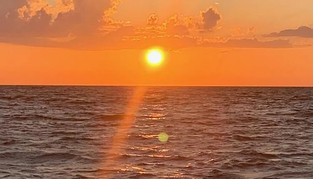 The image captures a serene sunset over the ocean with a vivid orange sky and the sun casting a reflection on the water