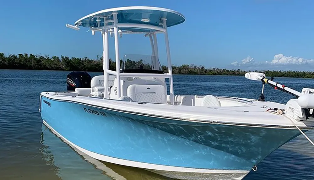 A center console boat painted in light blue and white is anchored near the shore under a clear sky