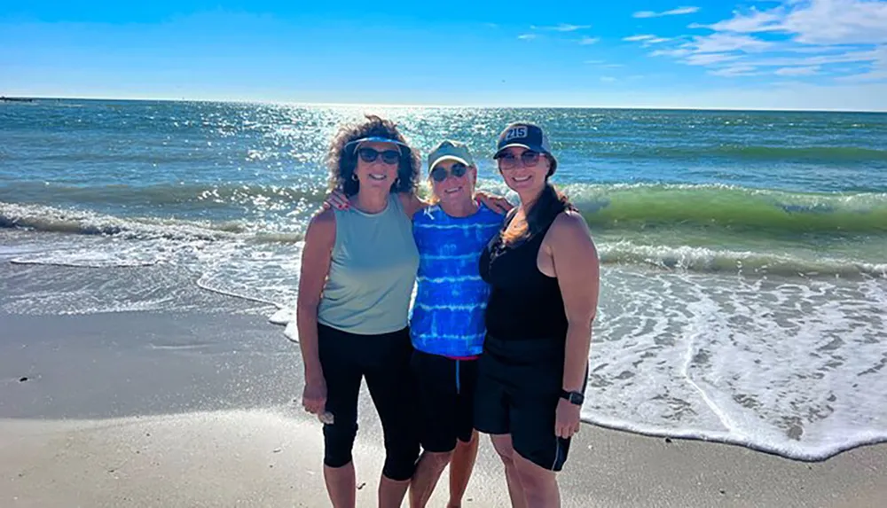 Three people are standing and smiling on a sunny beach with the ocean and waves in the background